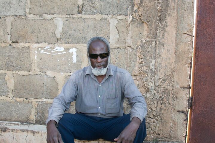 Elder in sunglasses seated against a block wall
