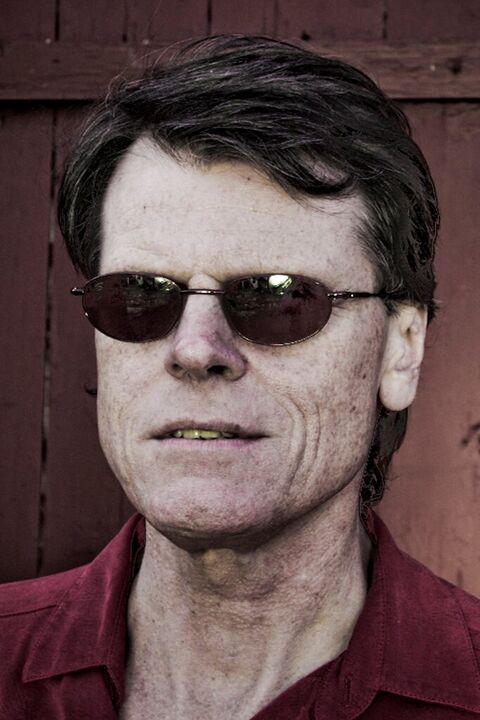 Man in dark sunglasses and a red shirt against weathered wood